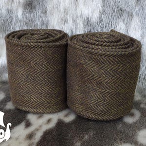 Könnte beinhalten: Zwei gerollte braune Stoffstücke mit Fischgrätmuster. Der Stoff ist eine Mischung aus braunen und goldenen Fäden, die ein strukturiertes Aussehen erzeugen. Die Rollen sind zylindrisch und stehen aufrecht, wodurch das Gewebe des Stoffes zur Geltung kommt. Der Hintergrund ist ein gesprenkeltes Grau und Weiß.