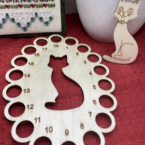 May include: A wooden cat-shaped thread organizer with 18 numbered holes. A separate wooden cat figurine is also present. The items are made of light-coloured wood and sit on a red surface. A cross-stitch project is in the background.