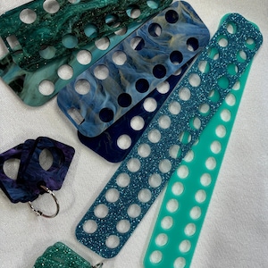 Peut inclure: Divers accessoires et porte-clés rectangulaires aux couleurs vives. Les articles présentent des nuances de bleu, vert et turquoise, certains avec des finitions pailletées. Chaque pièce est percée de multiples trous circulaires.