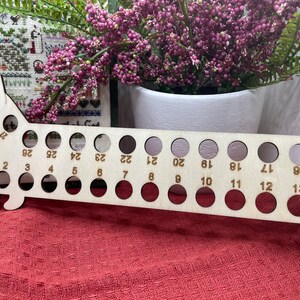 May include: A wooden dachshund-shaped needle gauge with numbered holes for measuring needle sizes. The gauge has 27 holes and is made of light-coloured wood. The gauge is on a red surface.