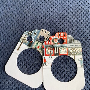 May include: Two white gift tags featuring a winter village scene. The tags have a round hole in the centre and depict red and blue buildings with snow on the ground. The text "Cafe Jolie" is visible on one of the tags.