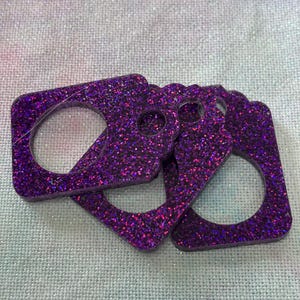 May include: Four purple glitter resin keyrings. Each keyring has a square shape with a rounded hole in the centre. The glitter is a mix of purple, pink, and black. The keyrings are on a light blue and white fabric.