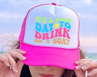 May include: Pink trucker hat with a white mesh back. The hat has a colorful graphic that says "It's a good day to drink on a boat".