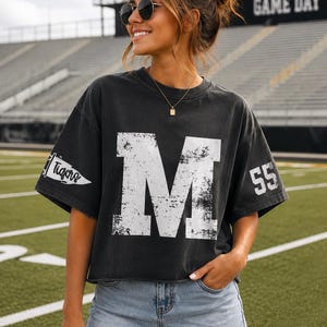 May include: A charcoal grey short-sleeve t-shirt featuring a large, distressed white letter M on the front, the word Tigers on a pennant graphic on the right sleeve, and the number 55 on the left sleeve, paired with light blue denim shorts.