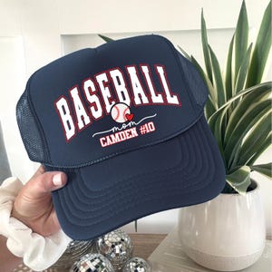 May include: Navy blue baseball cap with the word "BASEBALL" in white block letters. The cap features a baseball graphic with a heart and the text "mom CAMDEN #10". The cap has a mesh back.