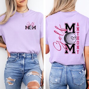 May include: A light purple t-shirt with the text "Golf Mom" and a golf ball graphic on the front. The back of the shirt features the text "Golf Mom Anthony" with a golf ball graphic. The shirt is made of a soft material.
