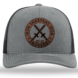 May include: A gray and black trucker hat with a brown circular patch featuring crossed swords and the text "IRON SHARPENS IRON 27:17 PROVERBS". The hat has a mesh back and a curved brim.