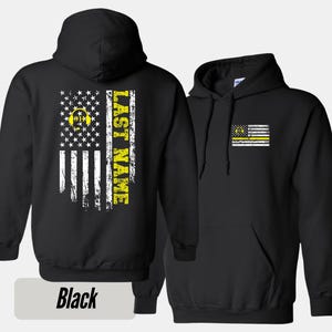 May include: Black hooded sweatshirt with a yellow and white American flag design on the back. The flag has a yellow graphic of a headset and the text "LAST NAME". The front of the sweatshirt has a smaller yellow and white American flag with a yellow graphic of a headset.