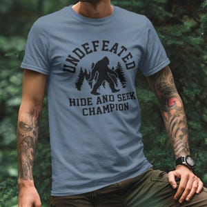 A man wearing a blue t-shirt with a graphic design of a Sasquatch and the text "UNDEFEATED HIDE AND SEEK CHAMPION" printed on it.