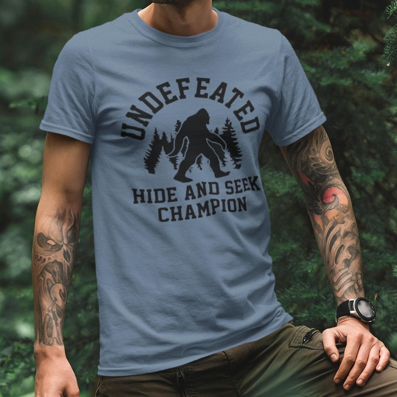 A man wearing a blue t-shirt with a graphic design of a Sasquatch and the text "UNDEFEATED HIDE AND SEEK CHAMPION" printed on it.