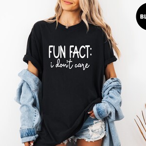 May include: Black t-shirt with the text "FUN FACT: i don't care" in white. The shirt is worn with a denim jacket and shorts. A black circle with the word "BLACK" is in the upper right corner.