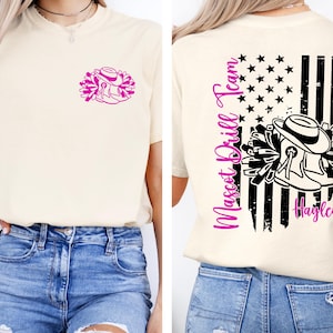 May include: Cream-colored t-shirt featuring a pink graphic of a cowboy hat, boots, and pom-poms. The back of the shirt displays an American flag design with the words "Mascot Drill Team" and the name "Hayley" in pink.