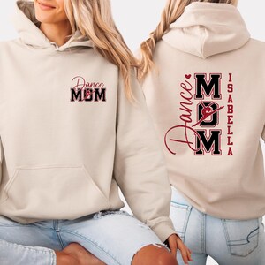 May include: Cream-coloured hoodie with a front pocket and a hood. The front features the text "Dance MOM" in a red and black design. The back of the hoodie displays "Dance MOM" with a dancer graphic, the name "Isabella", and a heart.