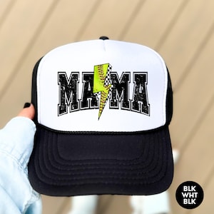 May include: A white and black trucker hat with a black and white chequered lightning bolt and a green softball graphic. The text "MAMA" is printed in black on the front of the hat.