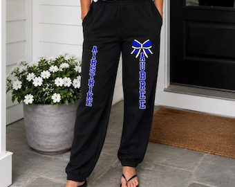 May include: Black sweatpants featuring a white and blue graphic of a bow above the text AUBREE and the vertical text AIRSTRIKE in blue block letters. The sweatpants have an elastic waistband and cuffed ankles, paired with a white t-shirt.