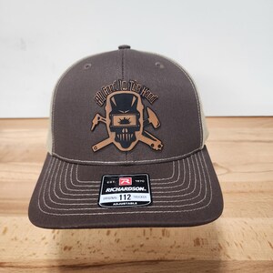 May include: Brown and beige trucker cap with a stitched brim. The cap features a leather patch with a skull wearing a welding mask and the text "All Food In The Hood". The Richardson brand label is visible.
