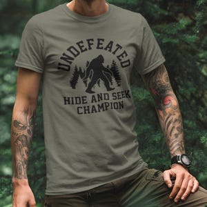 A person wearing a t-shirt with a graphic design of a Sasquatch silhouette and the text "UNDEFEATED HIDE AND SEEK CHAMPION" printed on it.