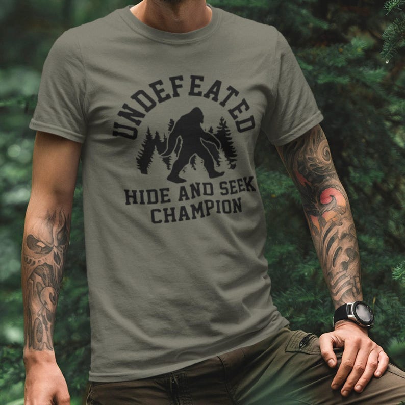 A person wearing a t-shirt with a graphic design of a Sasquatch silhouette and the text "UNDEFEATED HIDE AND SEEK CHAMPION" printed on it.