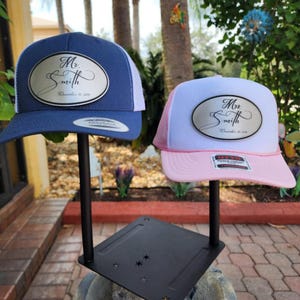 Peut inclure: Deux casquettes de camionneur avec des patchs ovales qui disent "Mr. Smith" et "Mrs. Smith" avec la date du mariage. Les casquettes sont exposées sur un support noir. La casquette de gauche est bleue avec un filet blanc à l'arrière. La casquette de droite est blanche avec un filet rose à l'arrière.