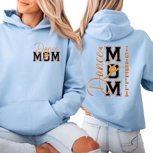 May include: Pale blue hoodie featuring the text "Dance MOM" in gold and black on the front. The back of the hoodie displays "Dance Mom Isabella" in gold lettering. The hoodie has a front pocket and a hood.