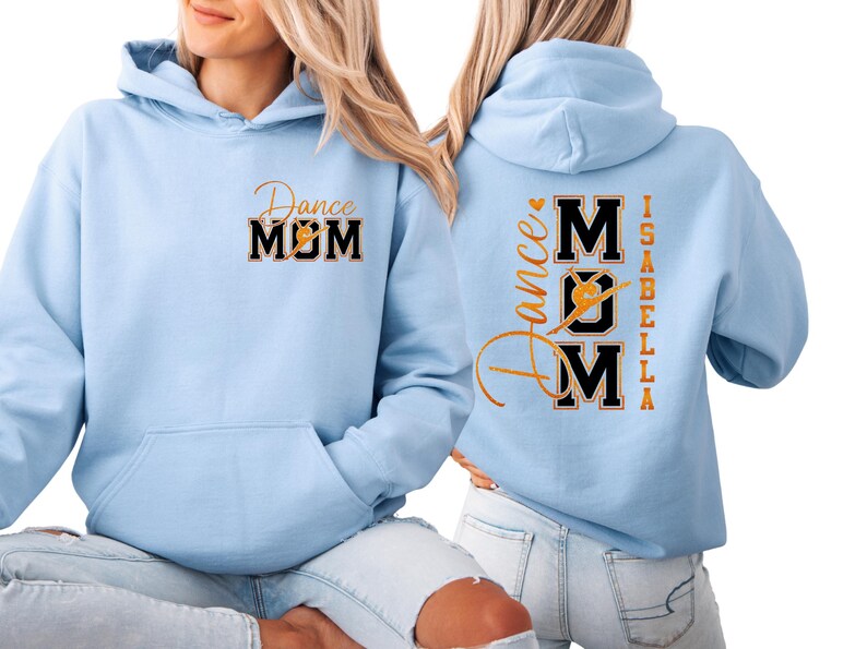 May include: Pale blue hoodie featuring the text "Dance MOM" in gold and black on the front. The back of the hoodie displays "Dance Mom Isabella" in gold lettering. The hoodie has a front pocket and a hood.