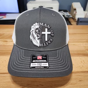 May include: A gray and white trucker hat with a black and white patch that says "Man of Faith" with a lion and a cross. The patch also includes the text "I Corinthians 16:13". The hat has a white mesh back and a black adjustable strap with a red and white logo that says "Richardson 112 Original Trucker Adjustable".