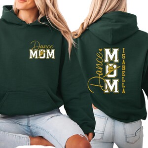 May include: A dark green hoodie with the words "Dance MOM" in gold script and white block letters on the front. The back of the hoodie features the words "Dance MOM" with a dancer graphic and the name "Isabella" in gold.