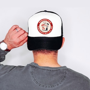 May include: A black and white trucker hat with a white front panel featuring a circular graphic. The graphic includes the text "REMEMBER KIDS ELECTRICITY WILL KILL YOU" around a cartoon image of a red devil and a person.
