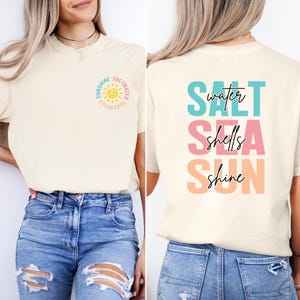 May include: Cream-colored t-shirt with a front graphic of a sun and the words "Sunshine, Saltwater, Seashells." The back of the shirt features the words "Salt, Water, Shells, Sea, Sun, Shine" in a stacked design with colorful fonts.