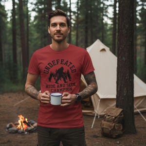 A man wearing a red t-shirt with a graphic of a Sasquatch and the text "UNDEFEATED HIDE AND SEEK CHAMPION" is standing in front of a tent and a campfire in a forest setting.