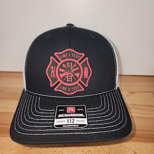 May include: Black and white trucker hat with a red firefighter emblem. The hat features a curved brim with white stitching and a white mesh back. The emblem has the text "LINE 1 TEXT" and "LINE 2 TEXT". The hat is adjustable.