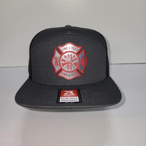 May include: A charcoal gray flat bill snapback hat with a mesh back. The front features a red and silver firefighter emblem with the text "LINE 1 TEXT" and "LINE 2 TEXT". The hat has a Richardson tag.