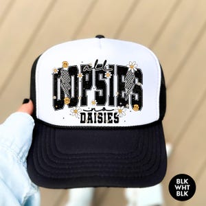 May include: A black and white trucker hat with a white front panel and a black mesh back. The front panel features a graphic of the text "a lil' OPSIES in DAISIES" in black with a yellow lightning bolt and daisy accents.