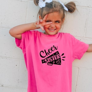 May include: A young girl wearing a bright pink t-shirt with the words "Cheer Sister" in black script and a megaphone graphic. She is smiling and making a peace sign with her fingers.