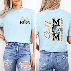 May include: Light blue t-shirt with "Golf Mom" printed on the front and back. The front features the text in black and gold with a golf ball graphic. The back has the text "Golf Mom" and "Anthony" with a golf ball graphic.
