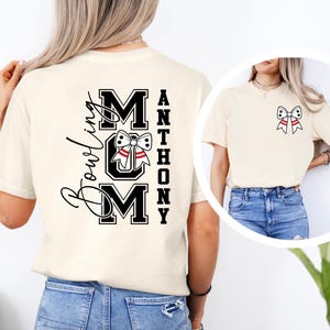 May include: Cream-coloured t-shirt with a bowling-themed design. The back features the words "Bowling Mom" and "MOM" in a bold, black font, with a bowling pin and bow tie graphic. The name "Anthony" is printed vertically. A smaller bow tie graphic is on the front.