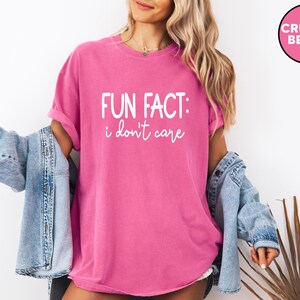 May include: A bright pink t-shirt with the text "FUN FACT: I don't care" in white script. The shirt is worn with a light blue denim jacket. A pink circle with the words "CRUNCH BERRY" is in the upper right corner.