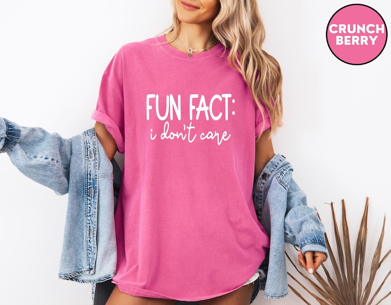 May include: A bright pink t-shirt with the text "FUN FACT: I don't care" in white script. The shirt is worn with a light blue denim jacket. A pink circle with the words "CRUNCH BERRY" is in the upper right corner.