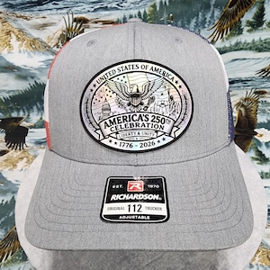May include: Gray trucker hat with a patriotic design. The front features a patch with an eagle, the text "United States of America", and "America's 250th Celebration 1776-2026". The hat has a mesh back and an adjustable strap.