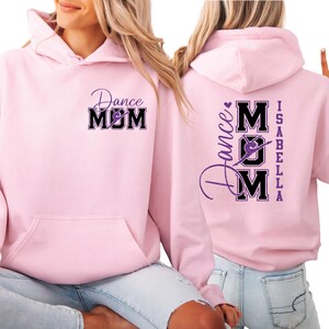 May include: Pale pink hoodie featuring the text "Dance Mom" in a black and purple design on the front and back. The back also has the name "Isabella". The hoodie has a front pocket and a hood.