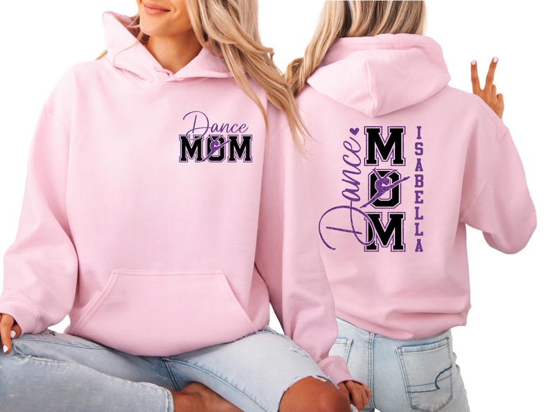 May include: Pale pink hoodie featuring the text "Dance Mom" in a black and purple design on the front and back. The back also has the name "Isabella". The hoodie has a front pocket and a hood.