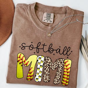 May include: A brown Comfort Colors t-shirt with the words "softball MIMI" in black script and block letters. The letters "MIMI" are designed with softball graphics and leopard print. A silver necklace and a brown hat are also visible.