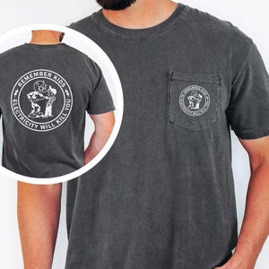 May include: Charcoal grey t-shirt with a pocket displaying a white circular graphic: "REMEMBER KIDS ELECTRICITY WILL KILL YOU." The same graphic is on the back. The shirt is short-sleeved.