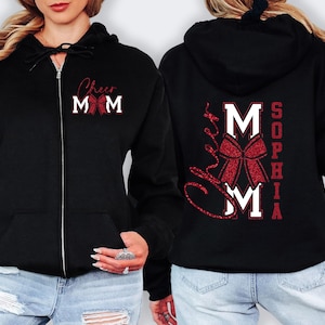 May include: Black zip-up hoodie with the text "Cheer Mom" and a red bow design on the front. The back of the hoodie features the name "Sophia" and the word "MOM" in a bold font with a red glitter effect. A stylish and comfortable garment.