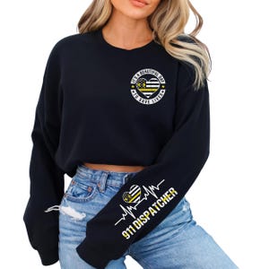 May include: Black cropped sweatshirt with long sleeves featuring a yellow and black heart with a thin yellow line flag design and the text "It's a beautiful day to save lives" on the front. The left sleeve has a yellow and black heart with a thin yellow line flag design and a heartbeat line with the text "911 Dispatcher" underneath.