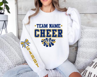 Peut inclure: Un sweat-shirt blanc à col rond avec l'inscription "TEAM NAME" et "CHEER" en lettres pailletées bleues et dorées sur la poitrine, accompagné d'un motif de nœud pailleté. La manche droite présente un nœud et le nom "GABBY" en lettres dorées.