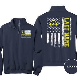 May include: A navy blue quarter-zip sweatshirt featuring a distressed American flag graphic. The front left chest displays a small flag with a yellow 911 headset icon. The back shows a large flag with the text LAST NAME in yellow.