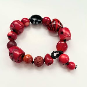 May include: A red coral beaded bracelet with a heart-shaped bead and a black bead. The bracelet is made with a variety of red coral beads, including some with a rough texture.