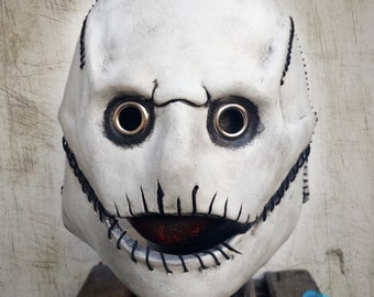 May include: A white leather mask with black stitching around the eyes and mouth. The mask has two large, black eye holes with silver grommets and a stitched mouth with a red interior.