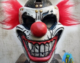 Puede incluir: Una máscara de payaso blanca y negra con una nariz roja y una amplia sonrisa de dientes. La máscara tiene un aspecto agrietado y desgastado, con pelo rojo a los lados. La máscara está sobre un soporte negro.